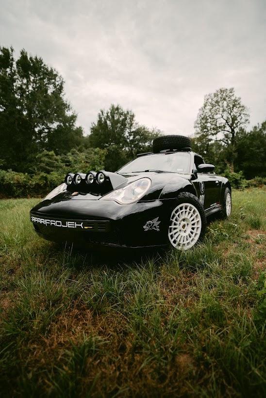 Black Porsche 911 Heritage RX with roof-mounted spare tire and hood light bar, parked in tall grass