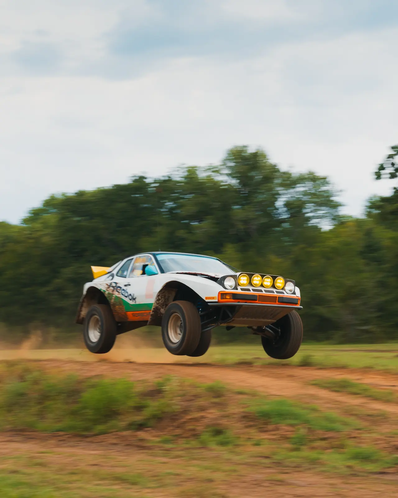 White and orange Porsche 944 rally car airborne over a dirt jump with all four wheels off the ground