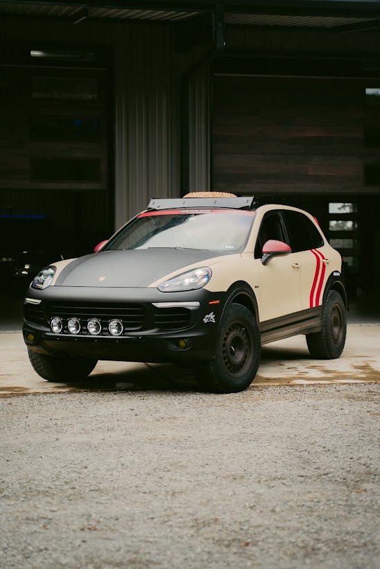 Tan and black Porsche Cayenne safari build with roof rack, steel wheels and front light pods, parked outside the shop